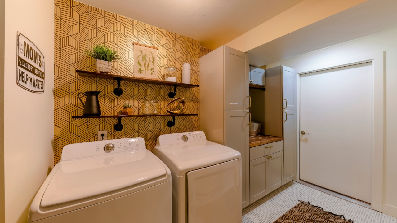 Decorative windowless laundry room with cabinets and counter. There are laundry machines against the gold wall paper with abstract pattern and shelves with displays beside the cabinets on the right.