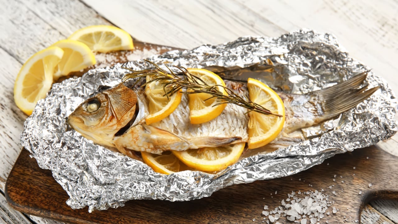 Aluminium foil with tasty baked fish, lemon slices and rosemary on light wooden background, closeup