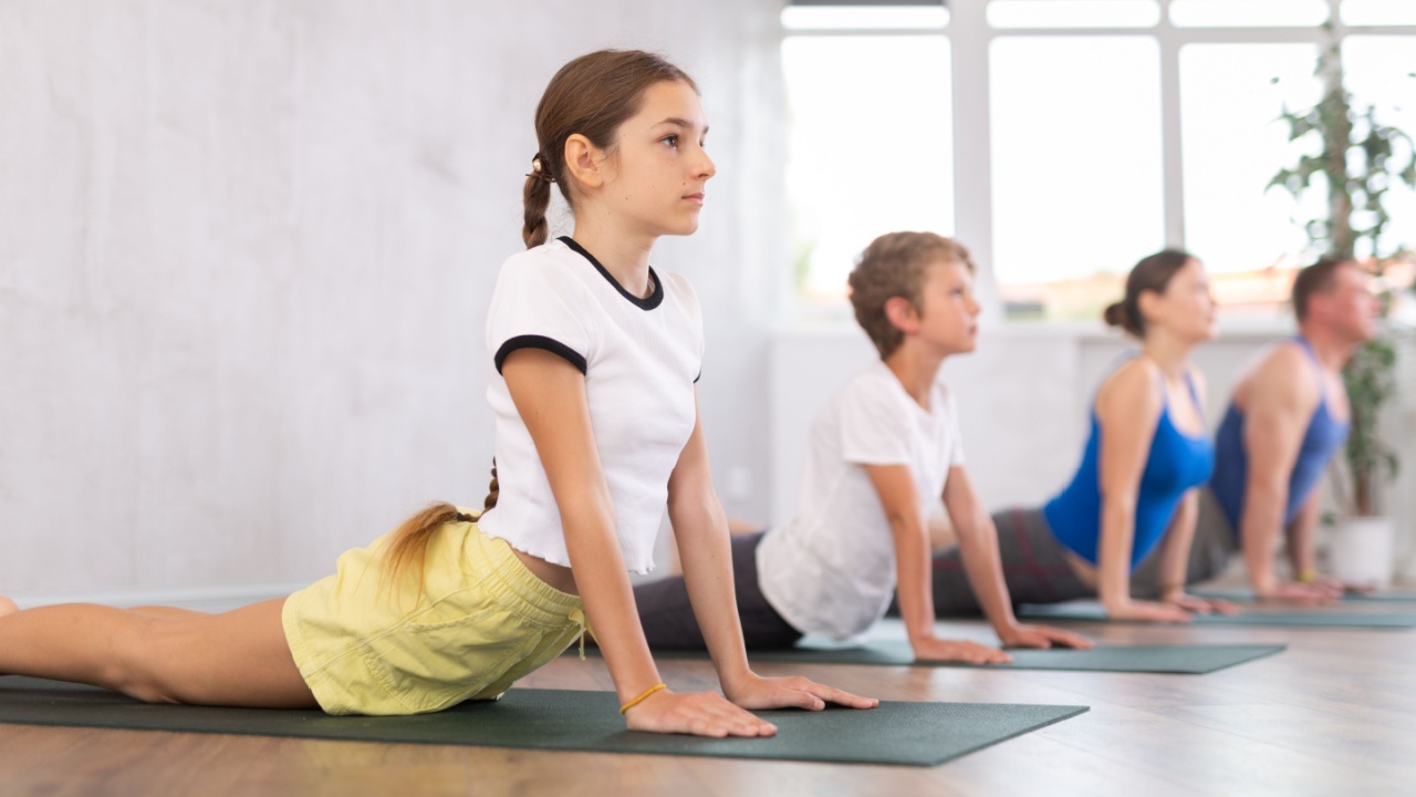 Friendly sporty parents with tween daughter and son performing Cobra Pose or Bhujangasana on gymnastic mats barefoot in yoga studio. Family health concept