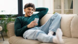 Mobile Offer. Smiling Middle Eastern Guy Browsing Internet On Smartphone, Communicating And Having Fun Online Lying On Couch At Home. Young Man Relaxing Using Gadget On Weekend