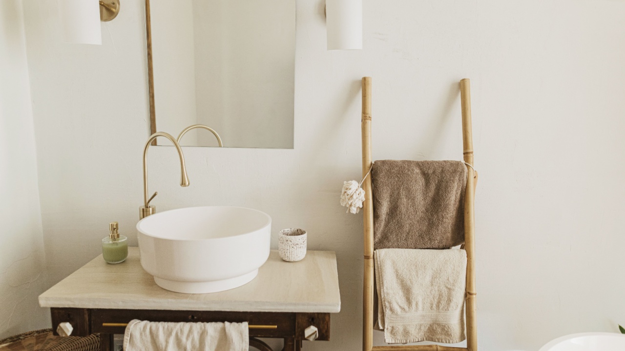 Stylish boho bathroom design. Modern ceramic sink with golden faucet on rustic stand with soap and ladder with towel on background of wall with mirror, modern bathroom interior.