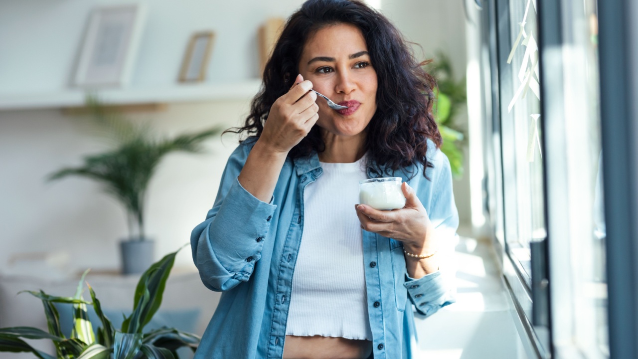 Shot of happy beautiful woman eating yogurt while standing in living room at home.