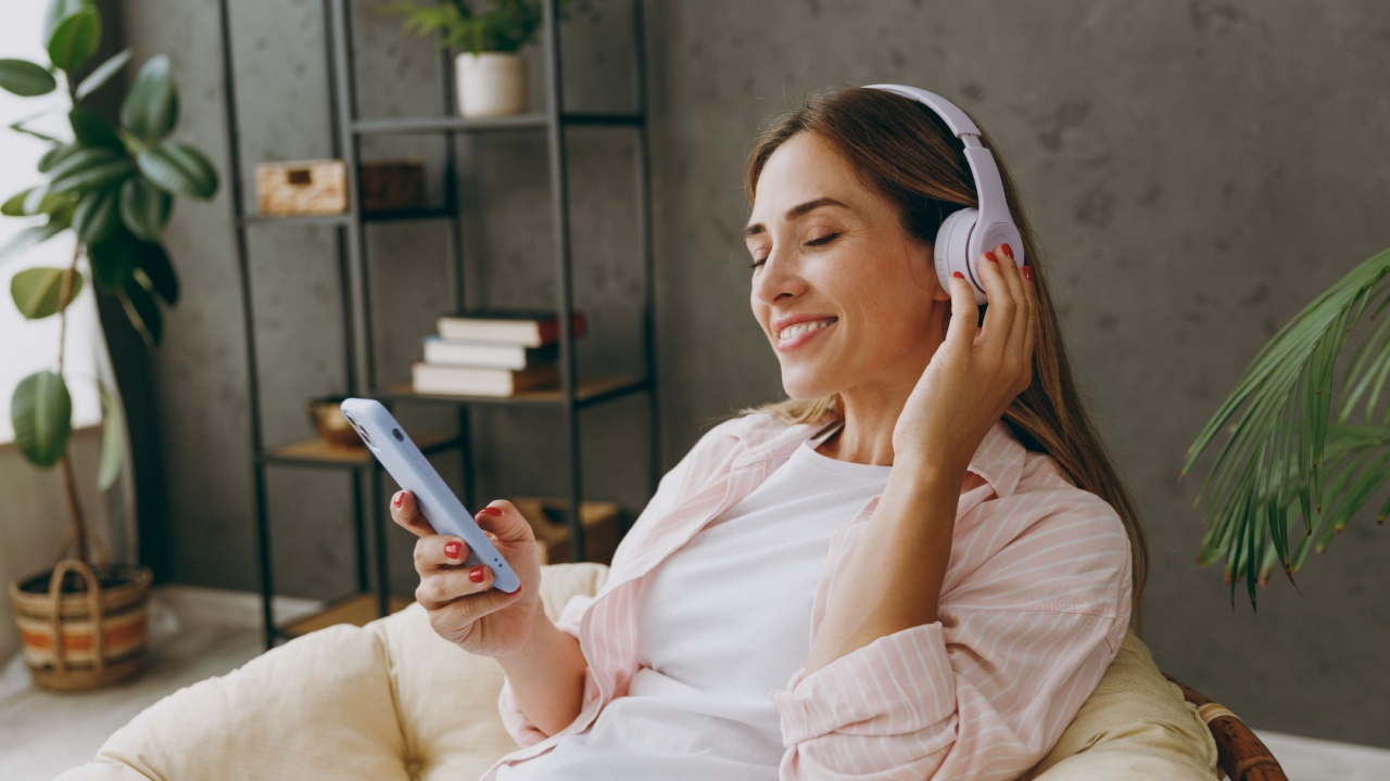 Young woman wear casual clothes sits in armchair listen to music in headphones use mobile cell phone stay at home hotel flat rest spend free spare time in living room indoor. Lifestyle lounge concept