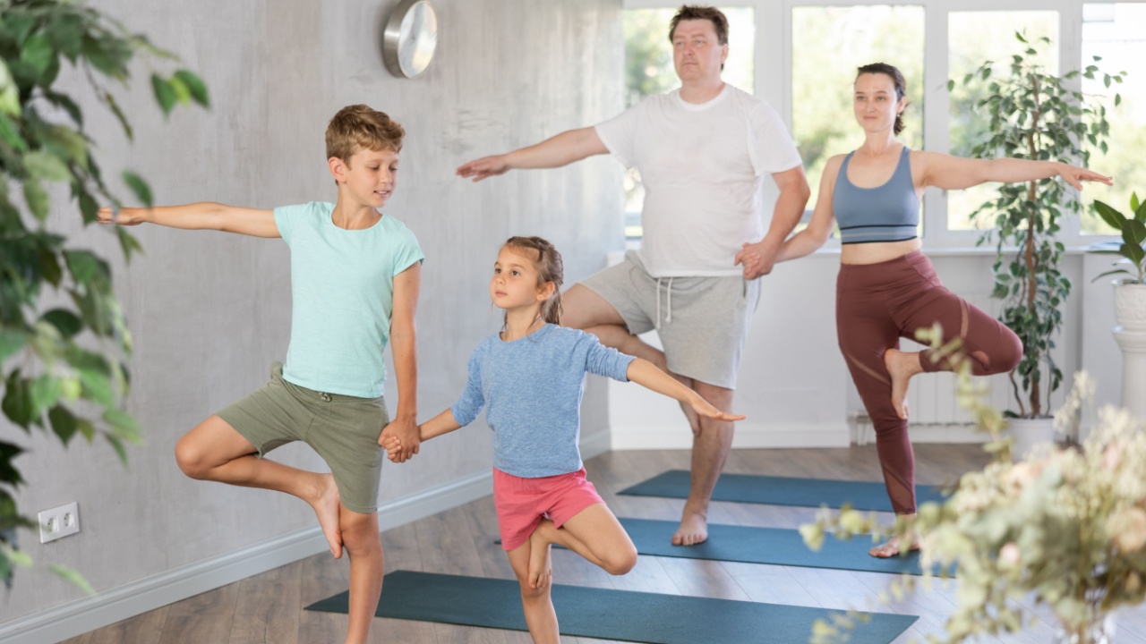 Sweet sister and brother practicing tree pose of yoga in pair together with their parents in light training room