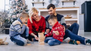 Happy young family with kids in woolen jumpers spending winter holidays at home playing with wooden blocks on warm floor in cozy, festively decorated living room with christmas tree