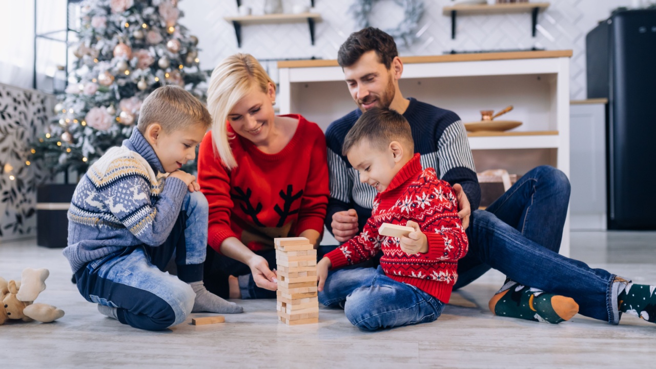 Happy young family with kids in woolen jumpers spending winter holidays at home playing with wooden blocks on warm floor in cozy, festively decorated living room with christmas tree