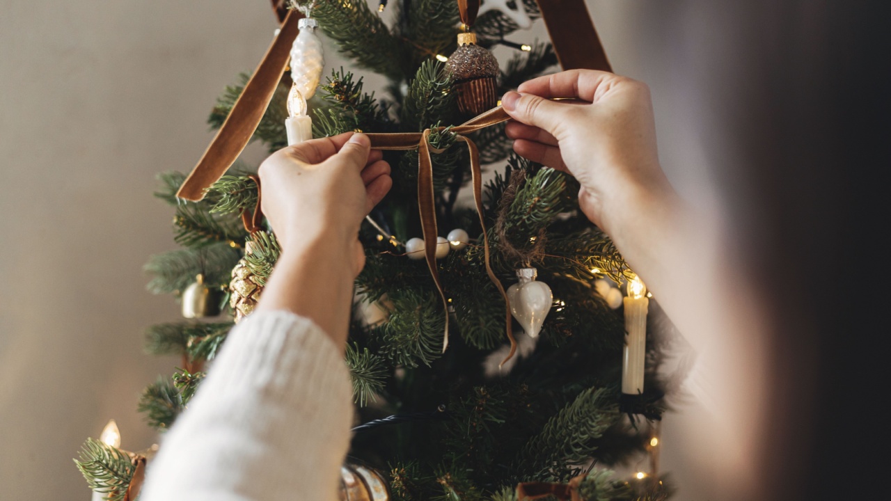 Hands in cozy sweater hanging modern velvet decoration on tree branch close up. Decorating christmas tree with stylish ribbon. Atmospheric winter holiday tradition, family time