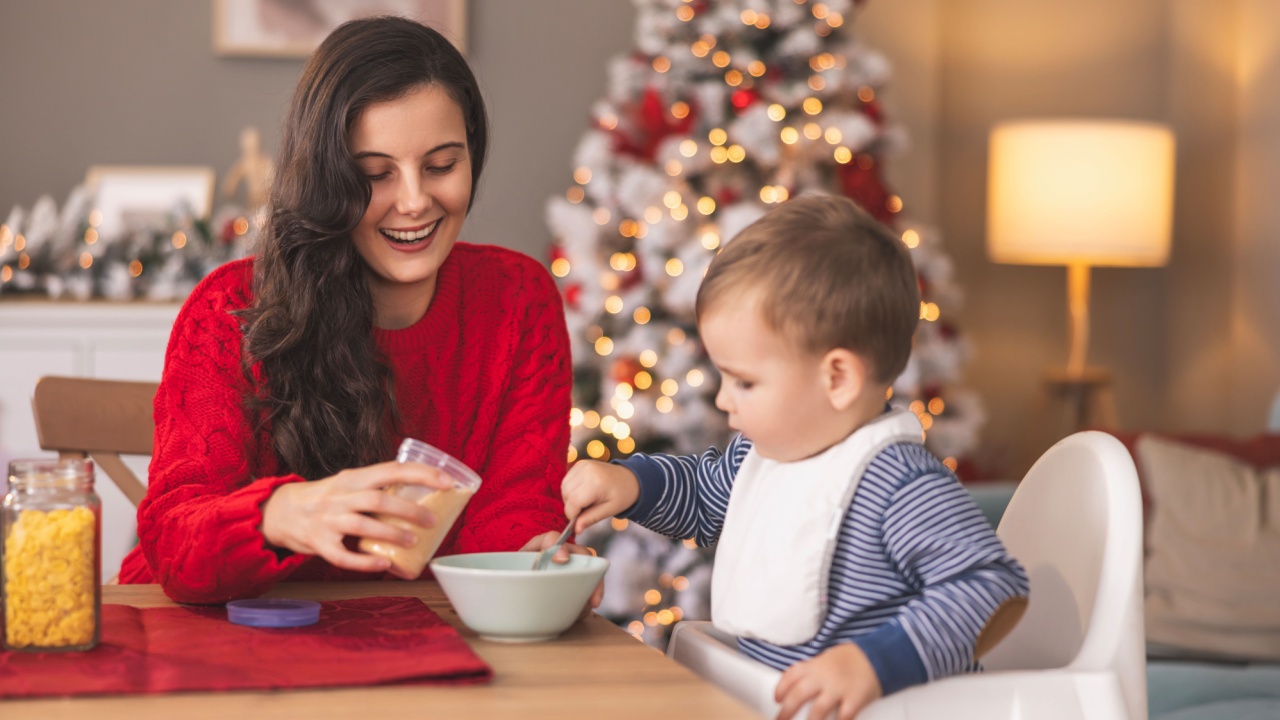 Cute toddler sittig in highchair playful while having breakfast on Christmas morning. Mother helping toddler boy while having breakfast on Christmas morning at home