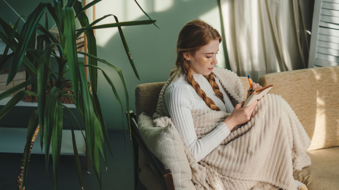Red haired woman studying from home in comfortable warm white winter blanket, writing ideas on a clipboard. Learning at home. Beautiful young woman sitting on
