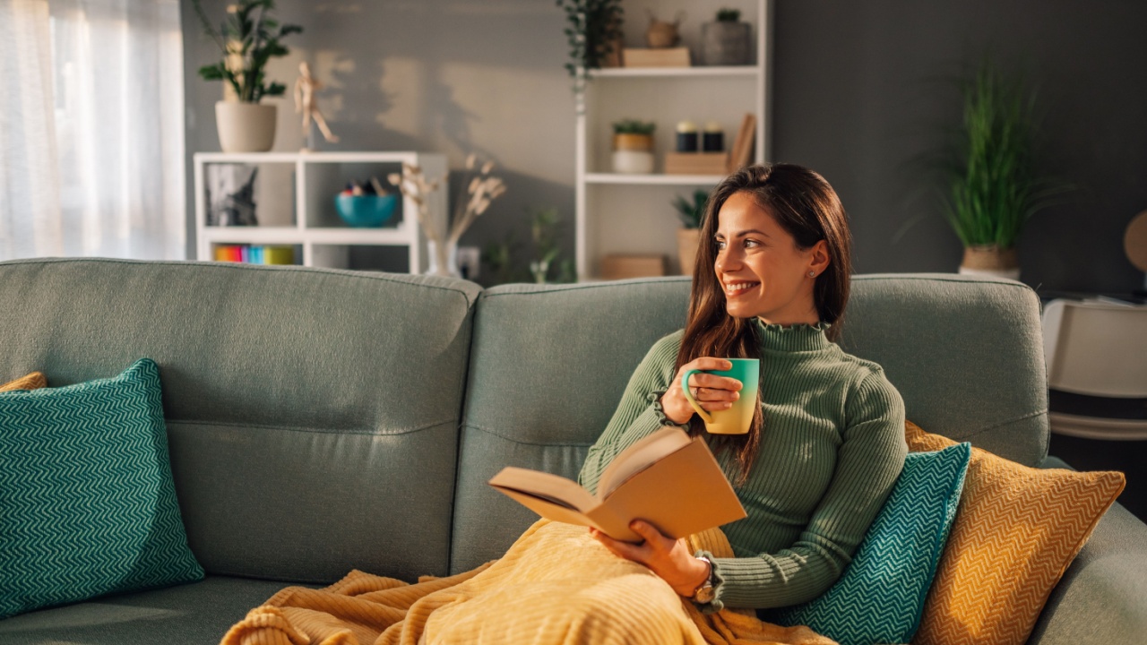 A young happy woman is relaxing at home while drinking coffee and reading a book. Relaxation at cozy apartment. A woman is sitting on sofa in a living room and enjoying weekend..