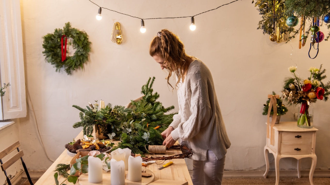 Young woman makes handmade Christmas wreath at home from natural materials. Authentic apartment with Scandinavian interior. DIY instruction from florist. Decorations with live pine needles. Copy space