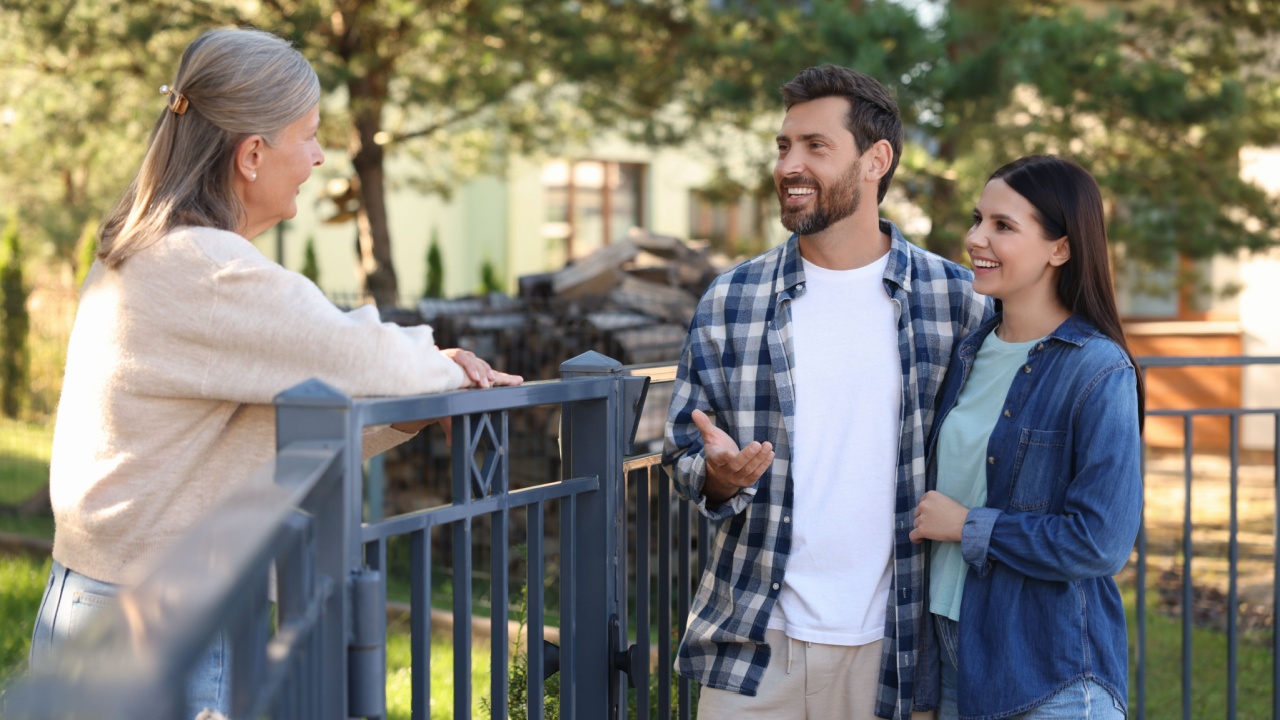 Friendly relationship with neighbours. Happy young couple talking to senior woman near fence outdoors