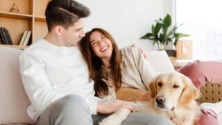 Portrait of attractive smiling young couple, boyfriend and girlfriend hugging, petting dog, golden retriever sitting on comfortable sofa in cozy home relaxing.