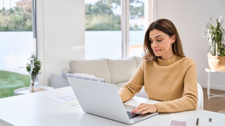 Smiling older mature middle aged woman using laptop computer sitting at table in living room, hybrid working, elearning with technology device at home, typing, browsing internet or searching online.