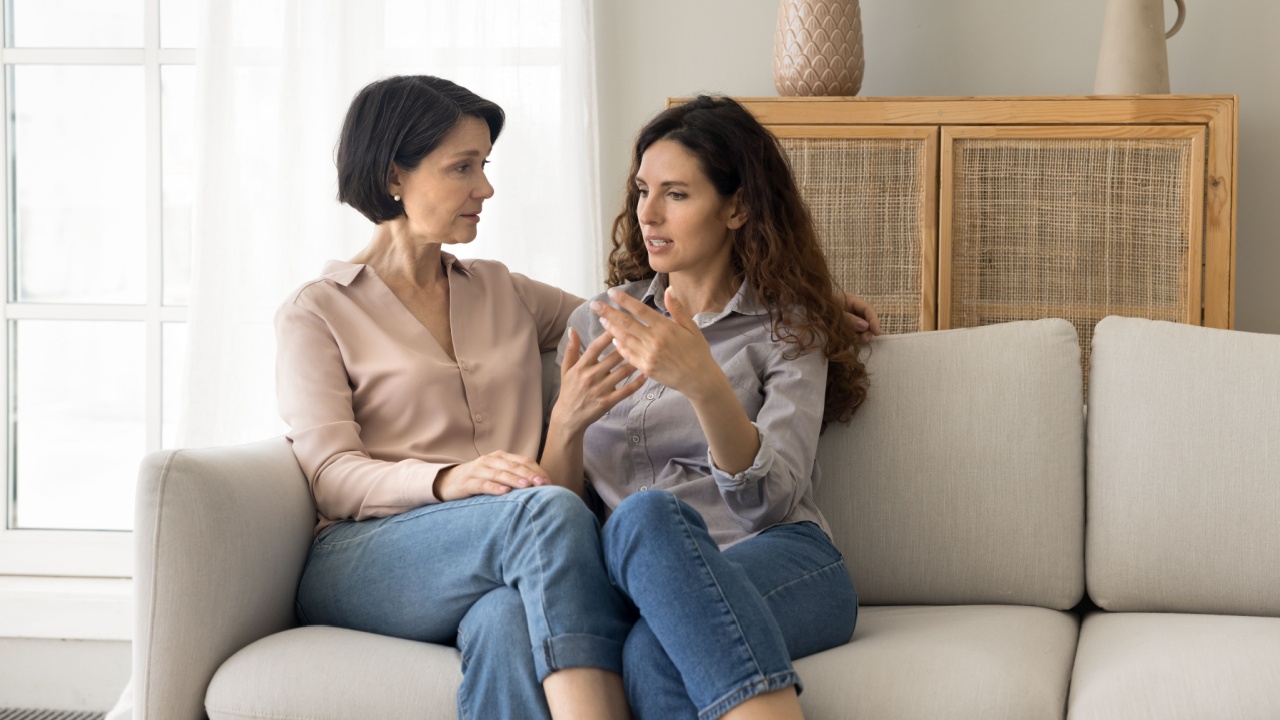 Attractive different age women, mature mom and young adult daughter lead trustworthy talk seated together on sofa, tell personal news, issues, receive support, asks advice, share life troubles at home