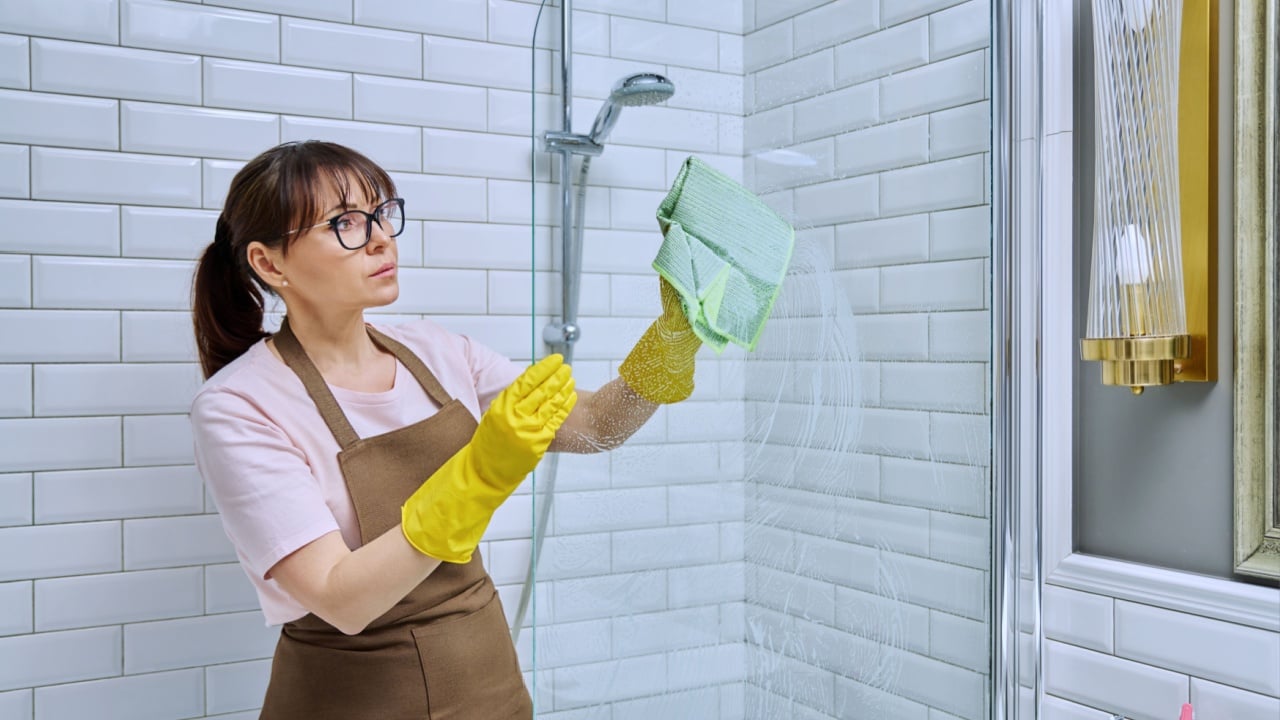Middle-aged woman in an apron cleaning glass in shower in bathroom