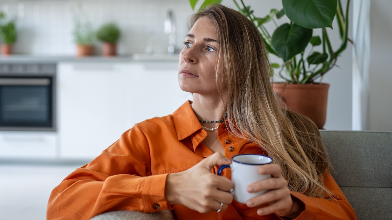 Thoughtful middle aged woman sits on sofa at home holding cup in hand looks towards window daydreaming. Peaceful pensive female thinking about future in cozy apartment in moment of quiet introspection