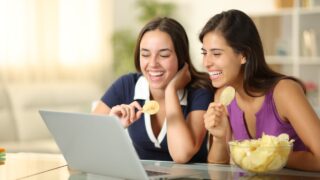 Happy women eating potato chips and watching video on laptop at home