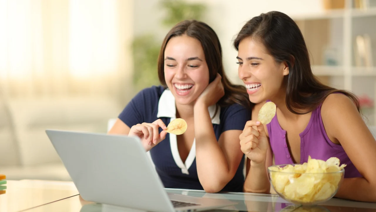 Happy women eating potato chips and watching video on laptop at home