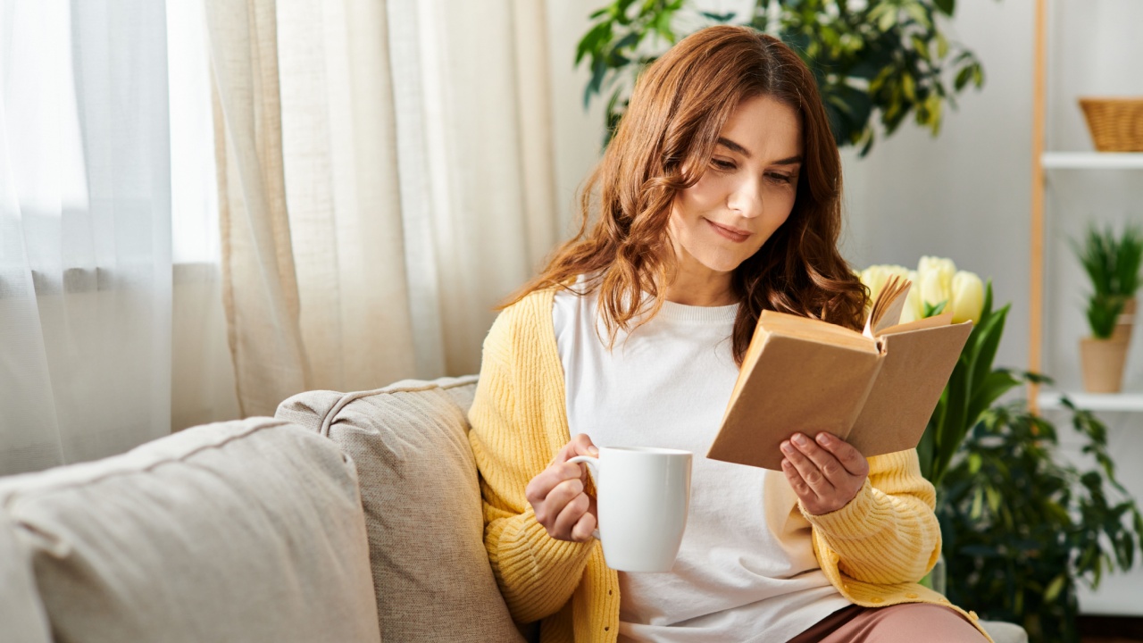 Middle-aged woman on sofa reads book, enjoying coffee.