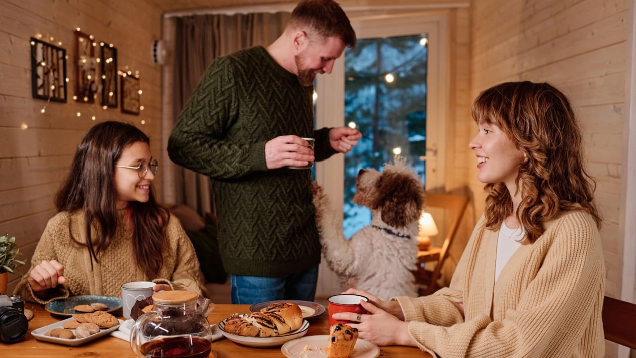Modern Caucasian family spending winter evening in wooden house drinking tea, chatting and playing with their dog