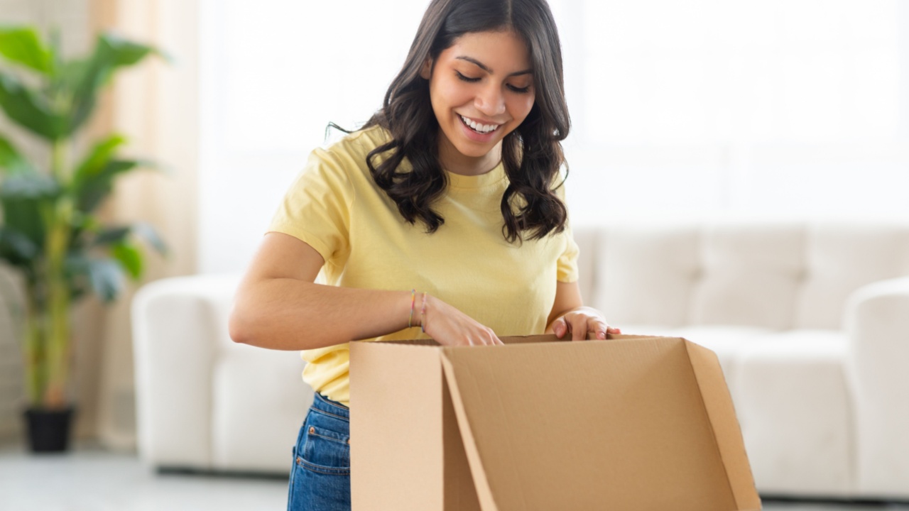 A middle eastern woman is standing at a table, with a box in front of her. She is in the process of opening the box, her hands carefully lifting the lid, looking inside