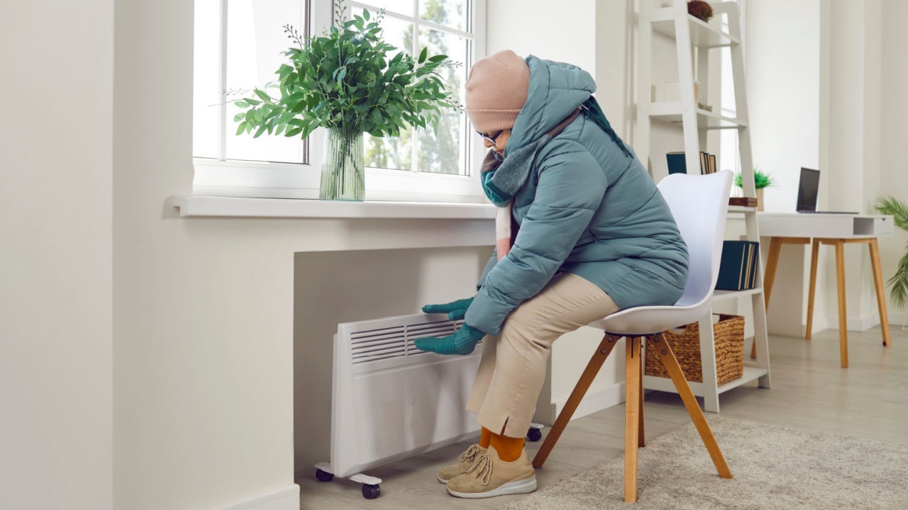 Elderly sad senior woman sitting on chair in winter outerwear in the living room warming up her hands on electric heater at home. Mature person freezing at home. Heating problems concept.