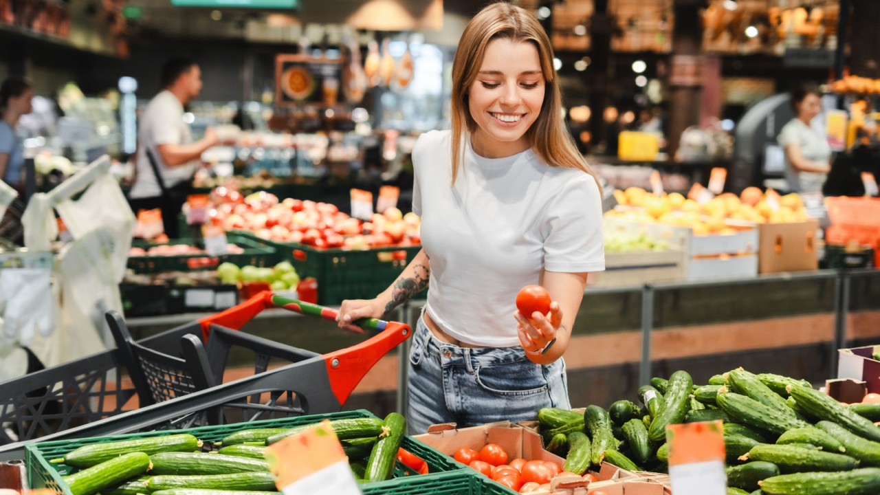 Young woman is smiling while choosing a tomato in the vegetable aisle of her local supermarket. She is pushing a shopping cart. Shopping concept