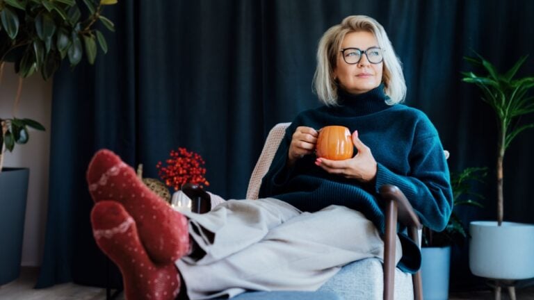 Middle aged woman relaxing with pumpkin shaped cup of hot drink in scandy style hygge interior home with fall mood decor. Lady dreaming, enjoy calm mood without stress, well being alone. Cozy autumn.