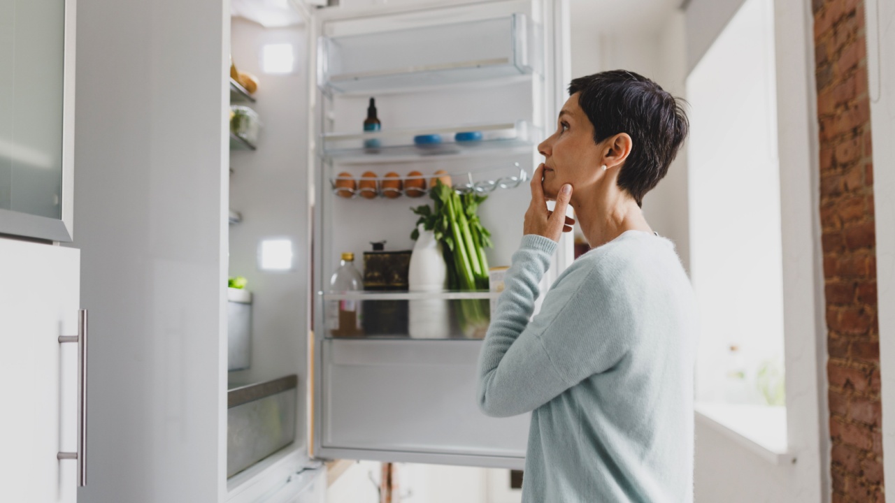 Side view of mature female with short hair standing in front of opened refrigerator at home with puzzled pensive facial expression, thinking of cooking breakfast, looking for ingredients