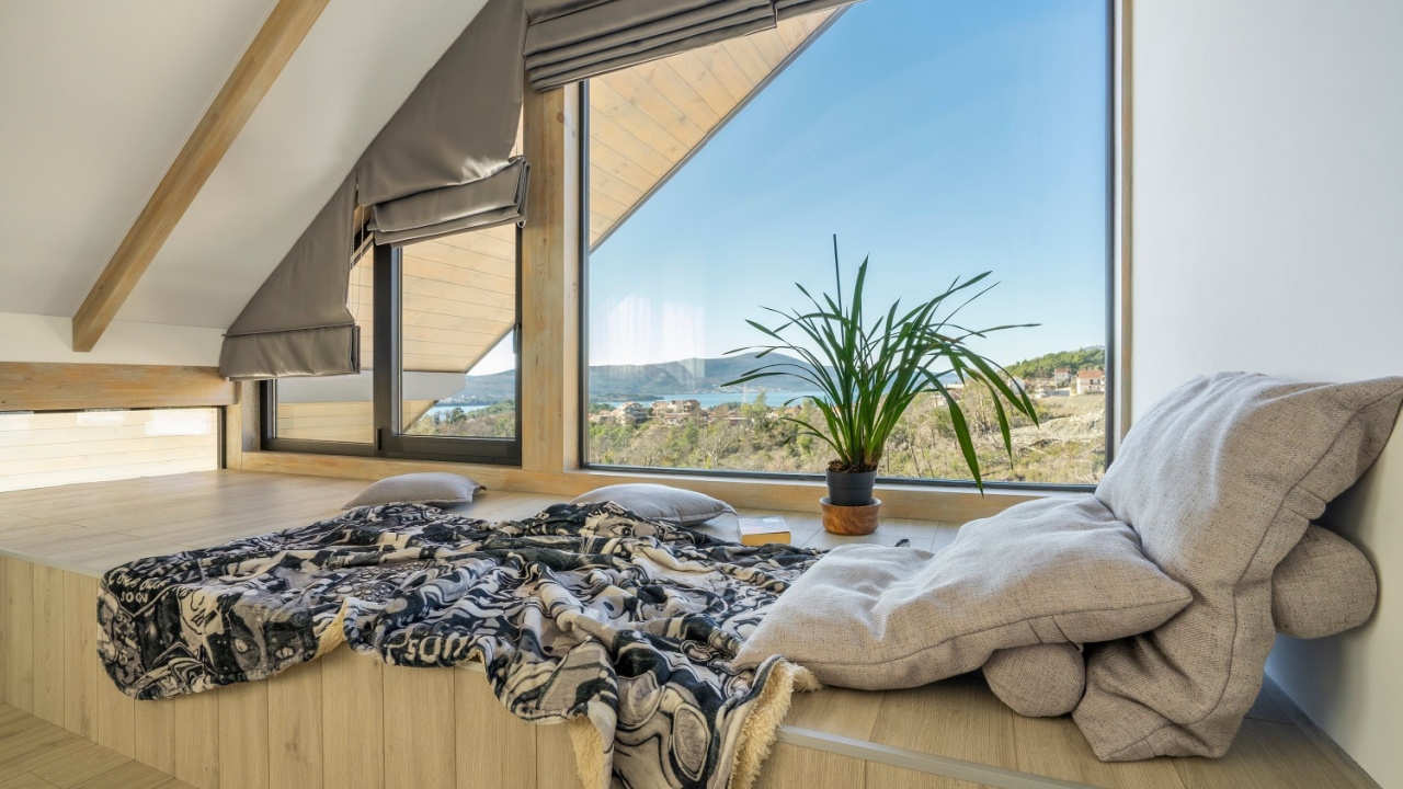 Cozy window nook with soft pillows and plaid invites relaxation. Natural light fills the space, showcasing a breathtaking view of a sea and hills through large windows. Sloped ceiling