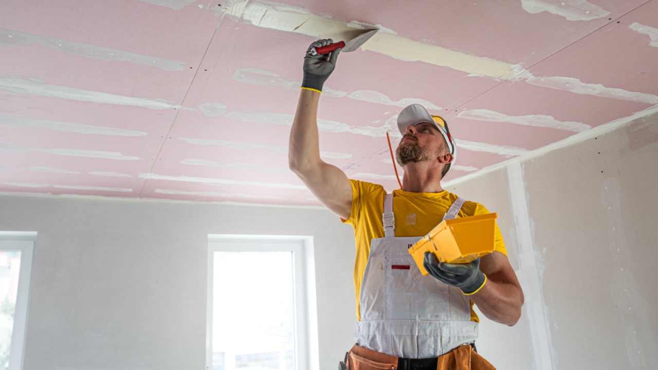 The worker make a plasterboard ceiling. He does taping plaster drywall ceiling joints. A red plasterboard improve the fire resistance of ceiling structures.