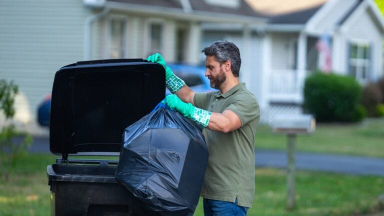 A man pushes a large plastic trash bin for weekly waste disposal schedule. Middle aged man putting out rubbish in garbage bin at the street. Man throwing trash bag into bin outdoors.