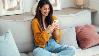 Shot of smiling young woman using her mobile phone sitting on couch at home