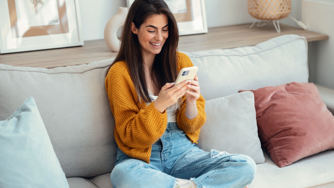 Shot of smiling young woman using her mobile phone sitting on couch at home