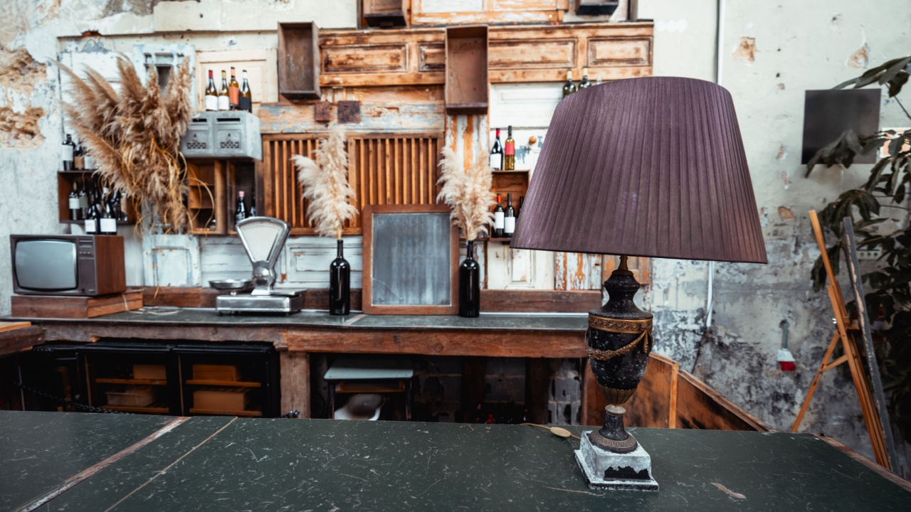 A stylish bar counter featuring a vintage table lamp with a pleated shade in the foreground. The background showcases rustic decor with wooden shelves, a wine rack, and pampas grass in vases