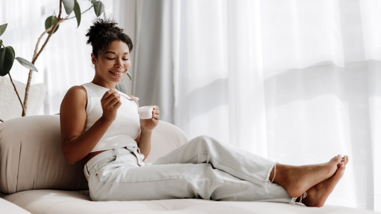 Young woman is relaxing on her couch at home, enjoying a cup of yogurt and smiling