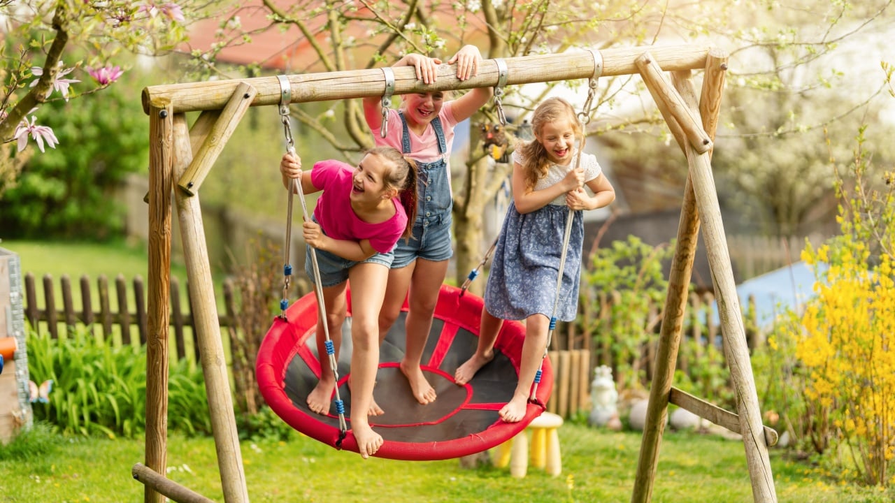 Three happy children are playing on a red round swing in a sunny backyard with blooming trees and a wooden fence, enjoying a fun moment outdoors.