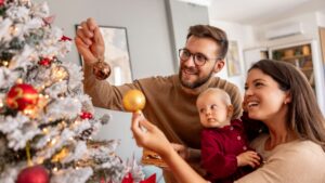 Young parents having fun decorating Christmas tree with their cute little baby girl, placing ornaments and decorating home for winter holiday season