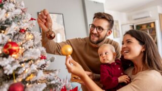 Young parents having fun decorating Christmas tree with their cute little baby girl, placing ornaments and decorating home for winter holiday season