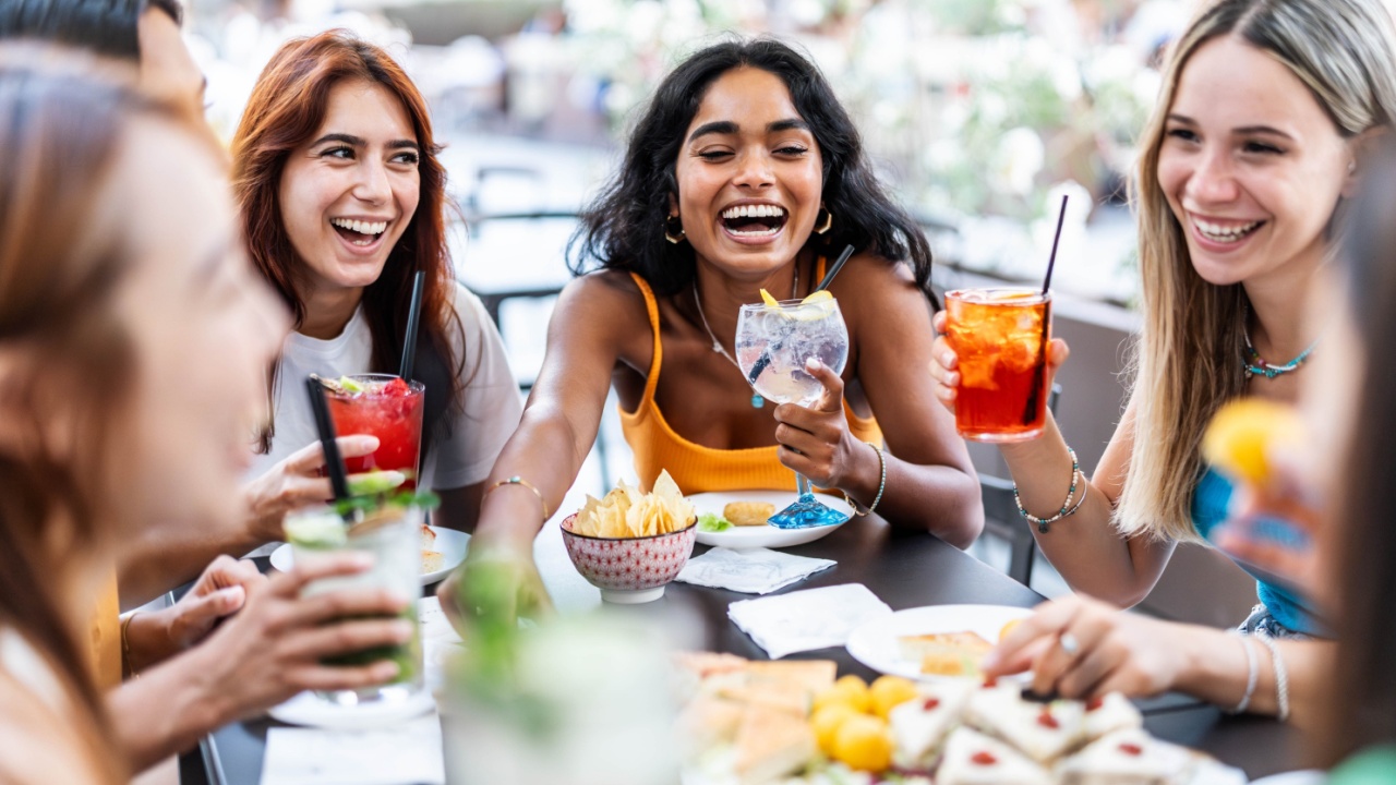 Happy female people toasting cocktail glasses sitting at bar table - Cheerful friends enjoying happy hour at pub restaurant balcony - Food and beverage life style concept