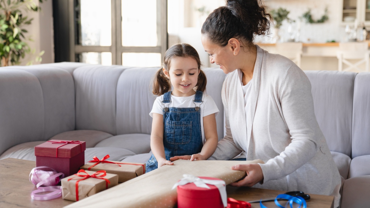 Little girl daughter helping her mom with wrapping packing in paper birthday Christmas New Year presents gift boxes at home together, preparing for celebration event