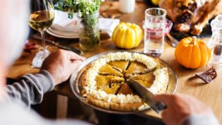 Pumpkin pie being sliced on a table with decorative pumpkins in Thanksgiving dinner. Warm autumn themed setting with people slicing pumpkin pie on wooden table. Thanksgiving pumpkin pie menu.