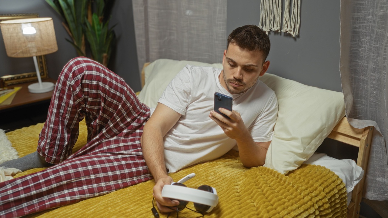 A young hispanic man with a beard is lounging on a bed in a bedroom, wearing pajamas, using a smartphone and resting a pair of headphones on a yellow blanket.