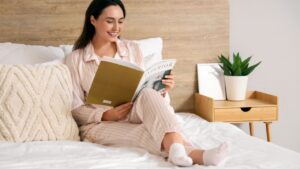 Young woman in pajamas reading magazine on bed at home