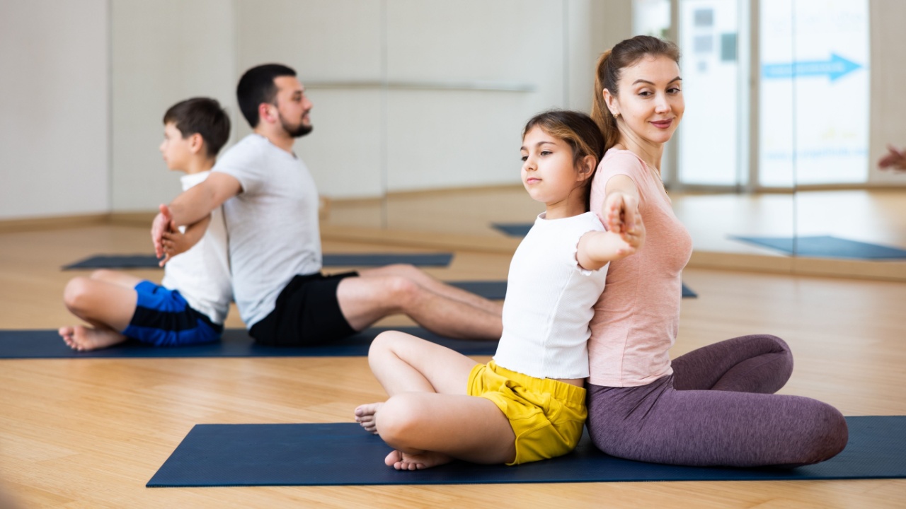 Kids and parents sitting back to back on mats during family yoga training in gym.