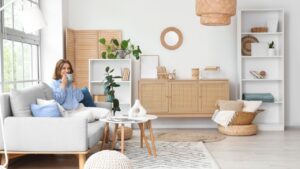 Pretty young woman with cup of coffee sitting on grey sofa near window in light living room