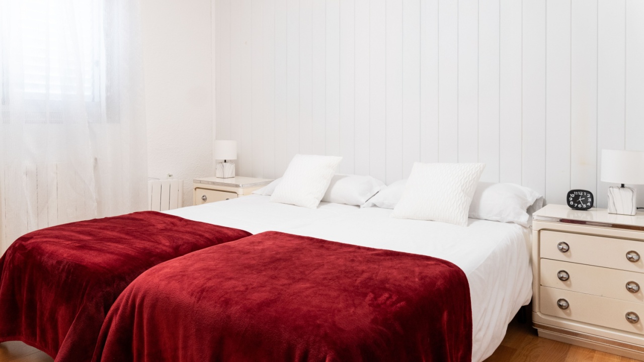 Cozy bedroom with white walls, wooden floor, and bedside tables featuring two single beds with red blankets