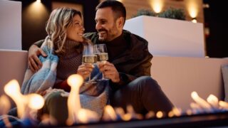 Happy couple toasting with wine while enjoying in romantic evening by fireplace on a patio.