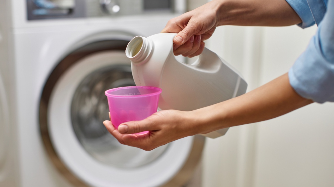 female hand pours liquid transparent laundry detergent or conditioner into plastic cap against blurred washing machine.